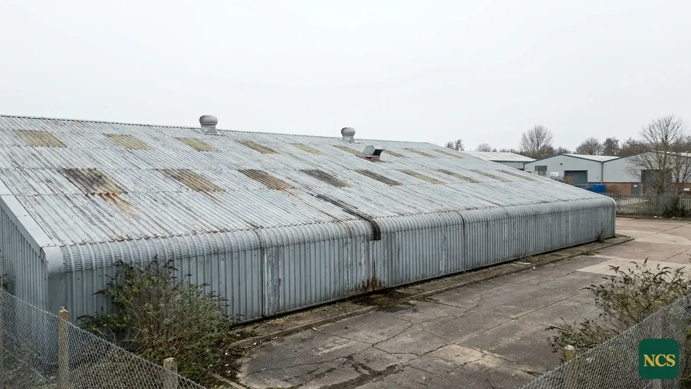 A weathered profiled steel commercial warehouse roof in overcast UK daylight, faded coating with patchy chalking and sur