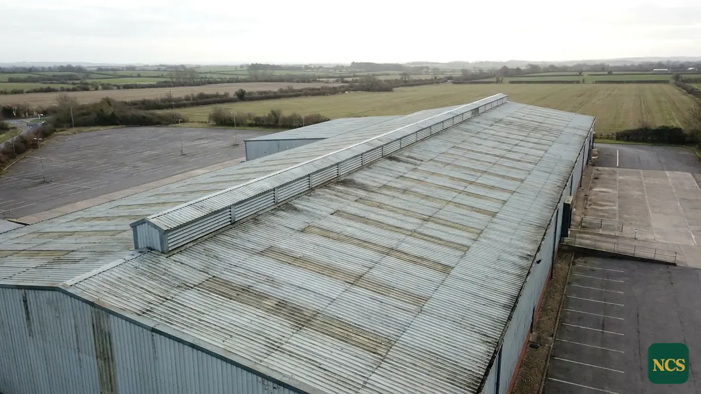 An exterior aerial view of a large profiled steel industrial warehouse roof in the UK under an overcast winter sky, fres